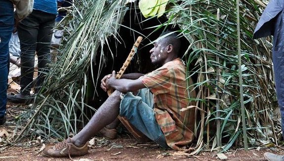 A member of the New Patrons of Cameroun is sitting on the ground between green branches