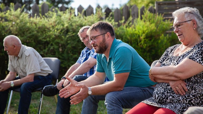 Members of the local council and local chief as New Patrons of Wietstock are sitting in a circle of chairs on a lawn during the presentation of the commission 