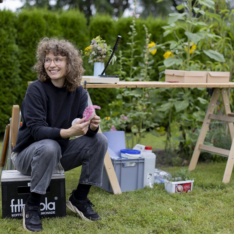 Die Neuen Auftraggeber von Wietstock Eine Person hockt auf einer Kiste im Garten und lacht