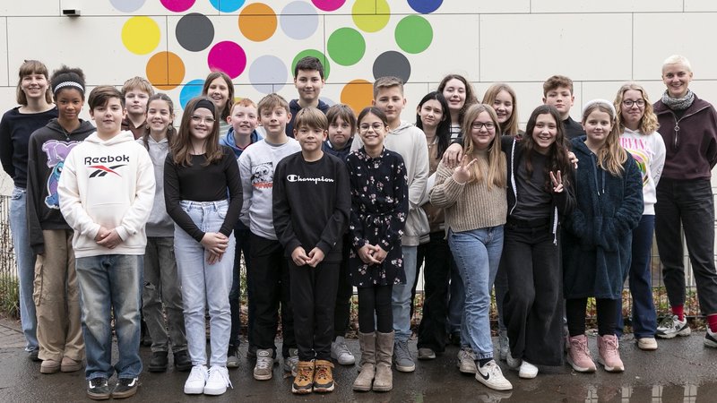 Gruppenbild von 23 Schüler*innen und zwei erwachsenen Frauen außen links und rechts, draußen vor einer Wand mit großen bunten Punkten