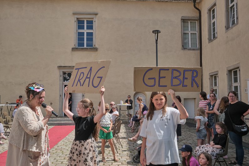 zwei Kinder halten Schilder mit "Trag" und "Geber" vor einem Publikum in einem Setting unter freiem Himmel. Neben ihnen steht eine Frau mit einem Mikrofon.