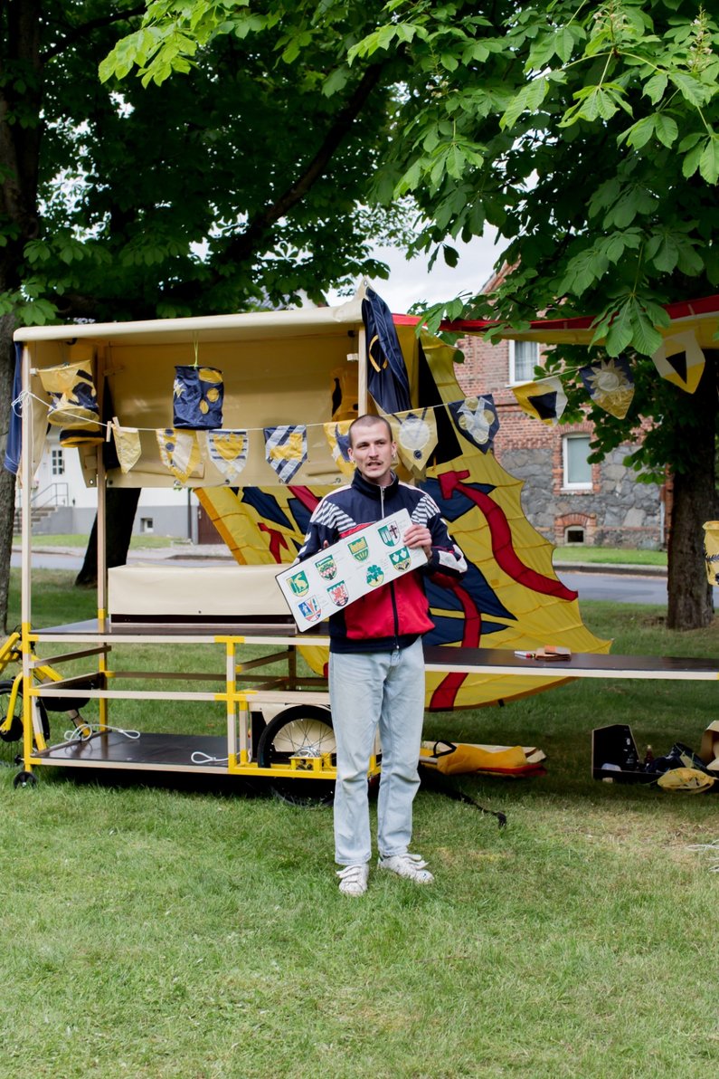 A person from The New Patrons of Steinhöfel stands in front of the Kistenflitzer holding village coat of arms in his hands