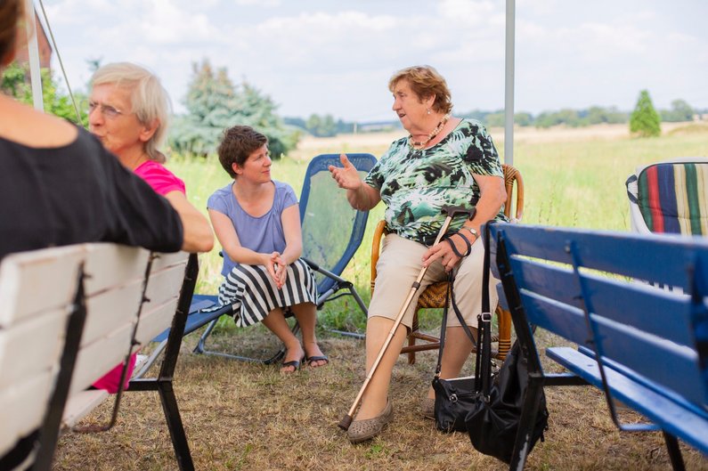 Koordinatorin Sophia Trollmann mit der Vorsitzenden des Seniorenbeirats Renate Kliems sitzen auf Bänken und diskutieren mit Personen der Neuen Auftraggeber von Steinhöfel