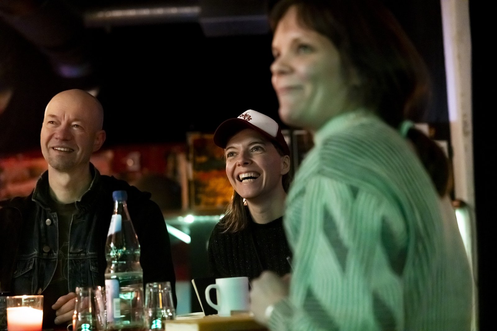 Three people are sitting at a table with drinks in a dark bar room. All three are looking in the same direction and laughing.