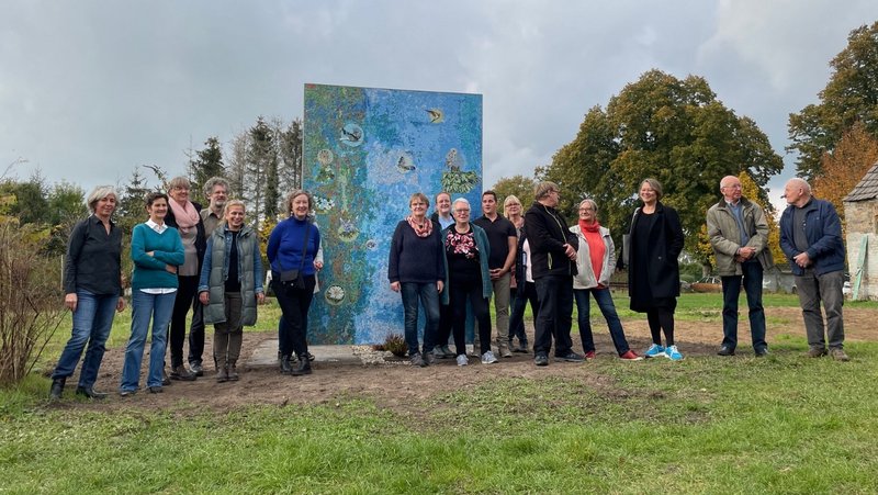 Group photo of The New Patrons' group in front of the mosaic based on the design "Animals and Plants in Wietstock" with artist Antje Majewski and mediator Susanne Burmester 