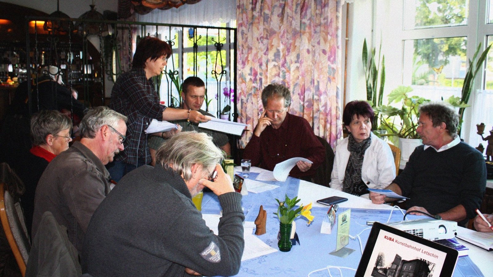 Members of The New Patrons of Letschin sit around a table and discussing