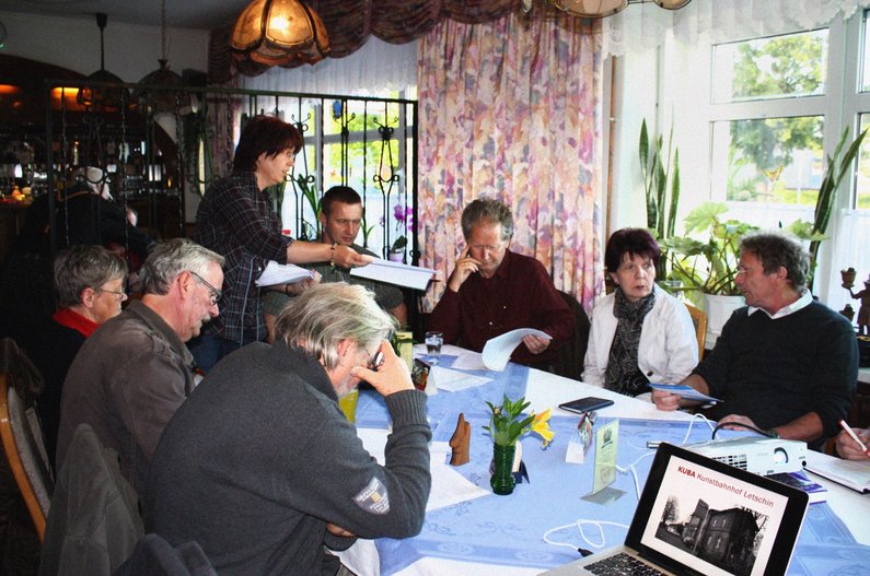 Members of The New Patrons of Letschin sit around a table and discussing