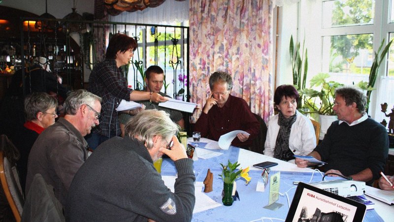 Members of The New Patrons of Letschin sit around a table and discussing