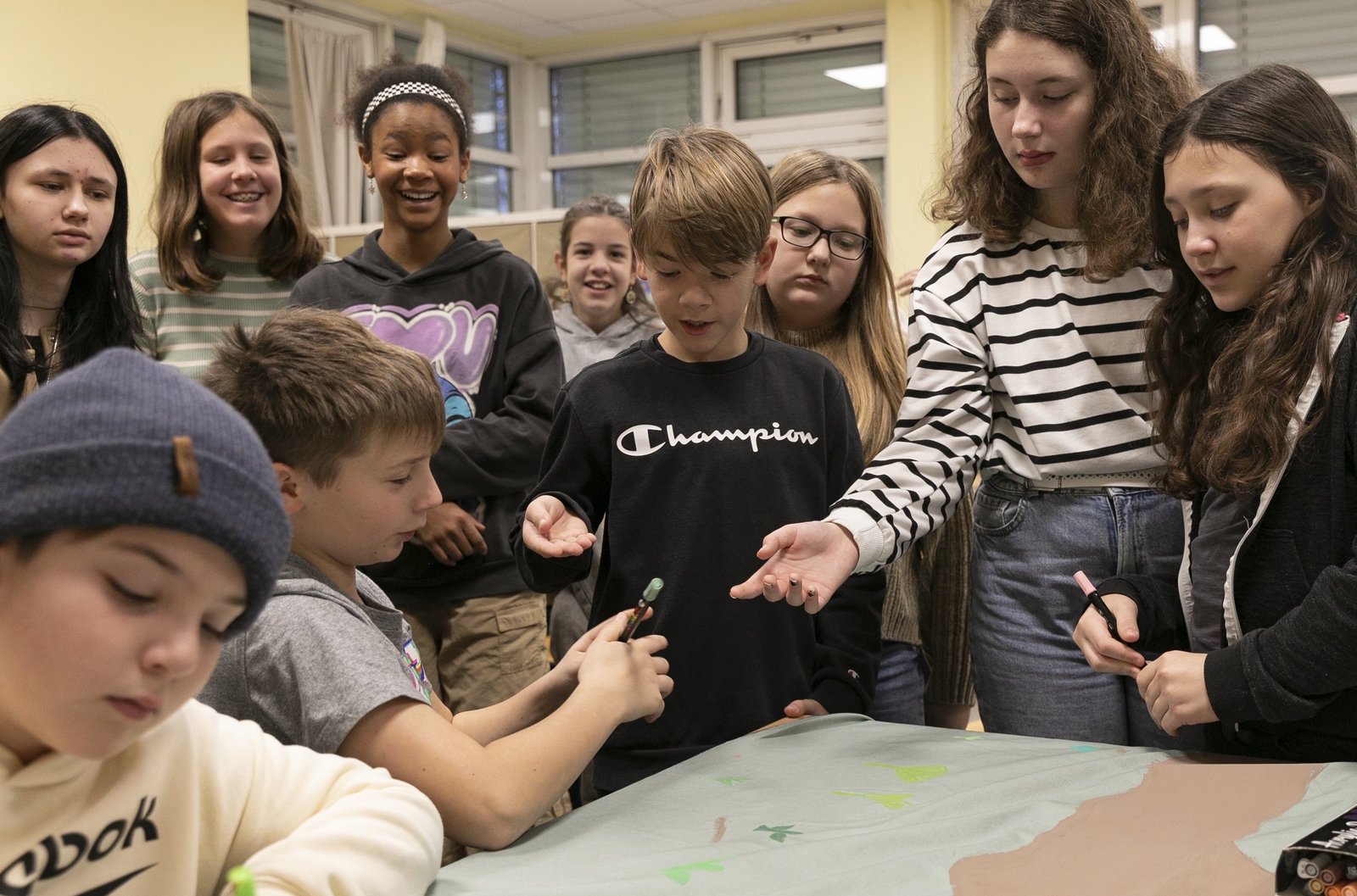A group of students sits and stands around a table with a piece of fabric with colored text on it.
