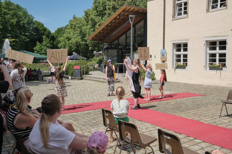 Drei Kinder stehen in einem Hof auf einem roten Teppich und halten Schilder hoch, auf denen zusammen "Neue Auftraggeber" steht. Weitere Kinder sitzen um den roten Teppich und schauen zu.