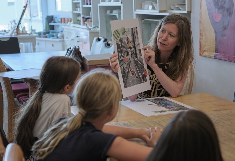 Eine Person mit langen blonden Haaren sitzt an einem Tisch mit drei Kindern und zeigt ihnen eine Collage.