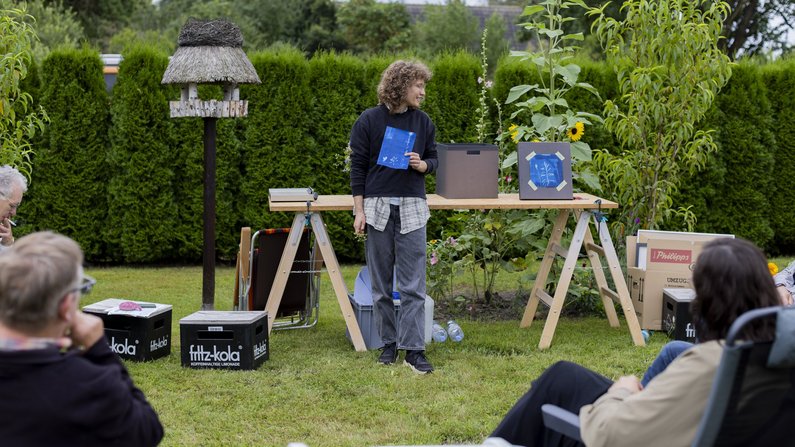 Die Neuen Auftraggeber von Wietstock Eine Person steht vor einem Tisch im Garten und zeigt ein Bild