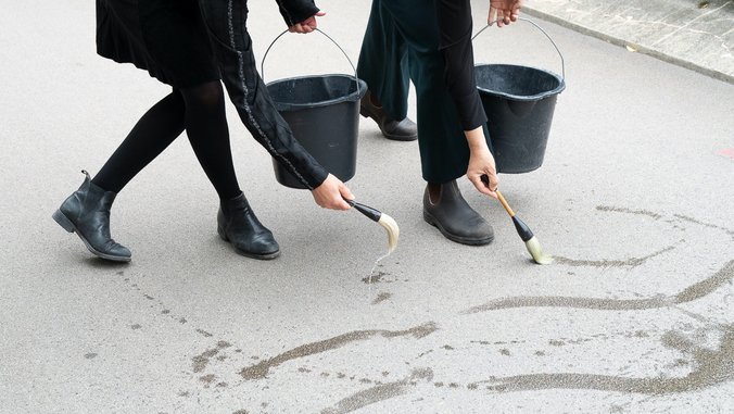 Auschnitt eines Bildes, die Hände von zwei Personen malen mit Pinseln auf Asphalt