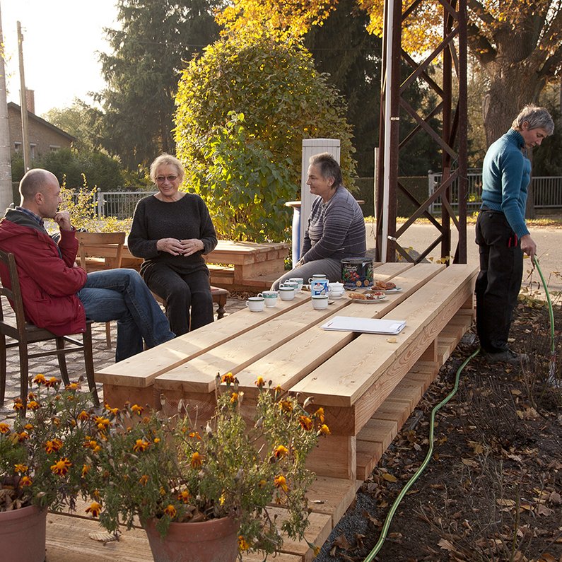 Vier Personen der Neuen Auftraggeber von Kleinliebenau sitzen draußen auf Stühlen und Holzsteg bei Café und Kuchen