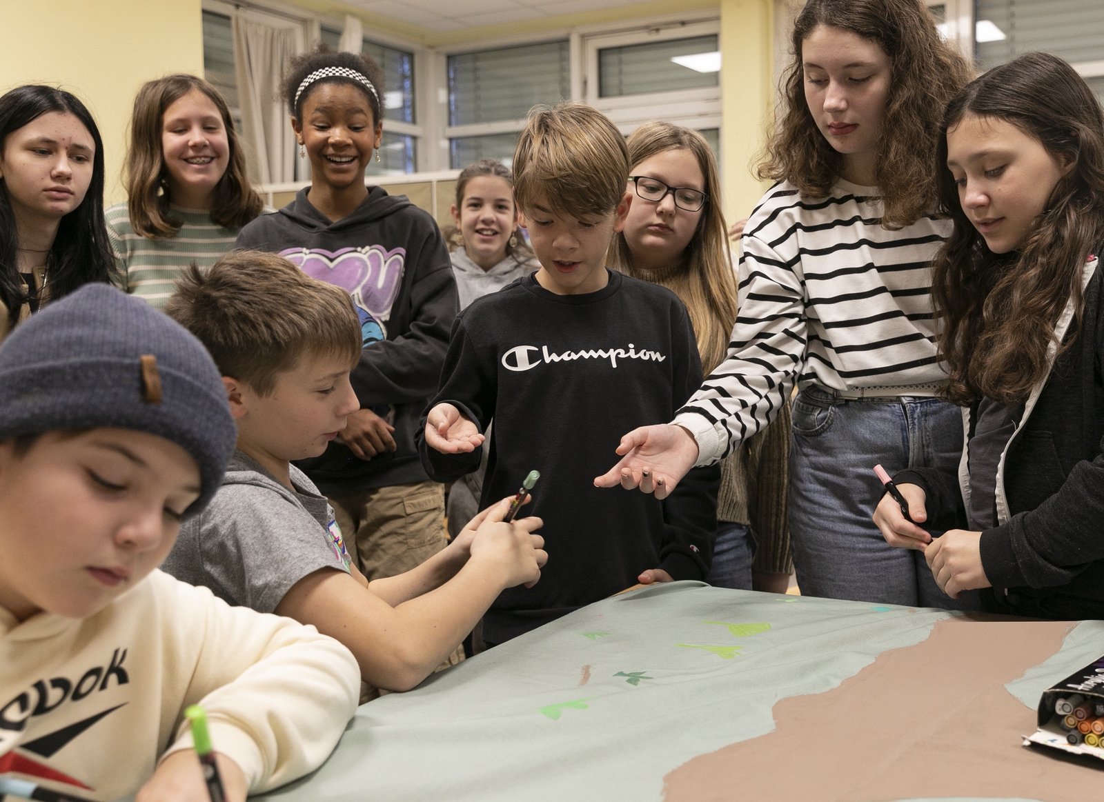 A group of students sits and stands around a table with a piece of fabric with colored text on it.