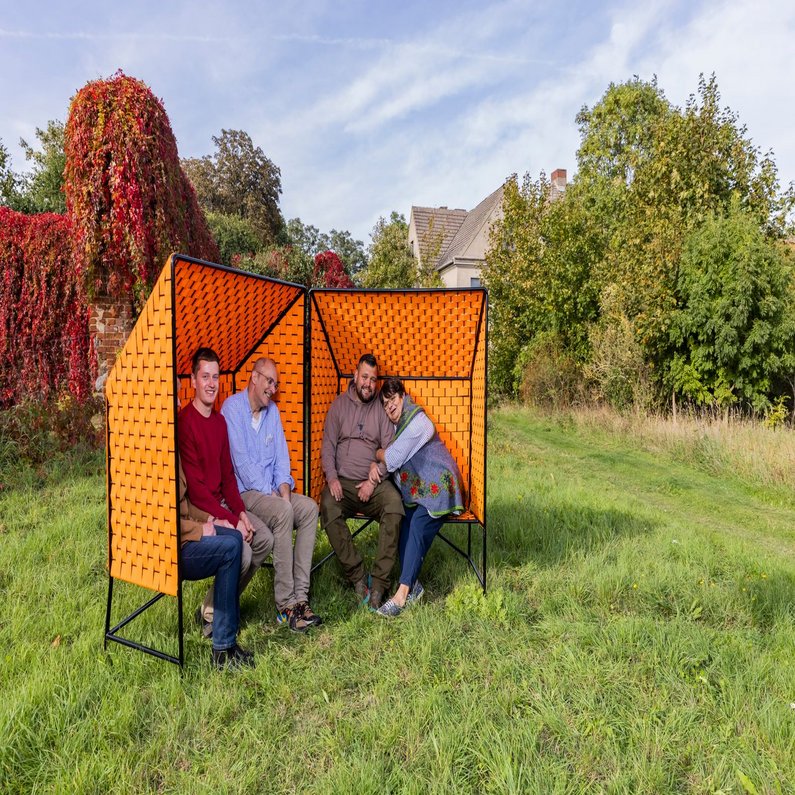 Photo shoot of the bank prototype The Züsedom-Bänke are standing in a field, members of The New Patrons and mediator Holger Friese are sitting in them and laughing.