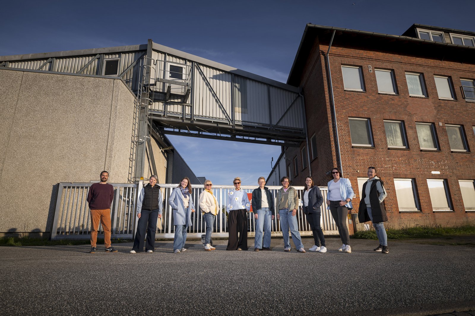 Eine Gruppe von menschen steht in einer reihe vor einem Fabrikgelände mit Backstein- und Betonbauten und einer Förderbrücke.