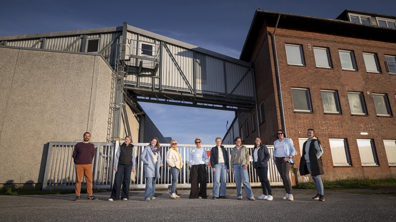 Eine Gruppe von menschen steht in einer reihe vor einem Fabrikgelände mit Backstein- und Betonbauten und einer Förderbrücke.
