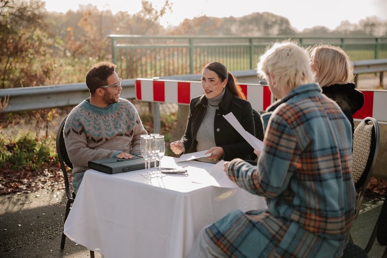 Vier Personen sitzen an einem Tisch im freien, gedeckt mit einer Tischdecke und Sekt, vor einer abgesperrten Brücke