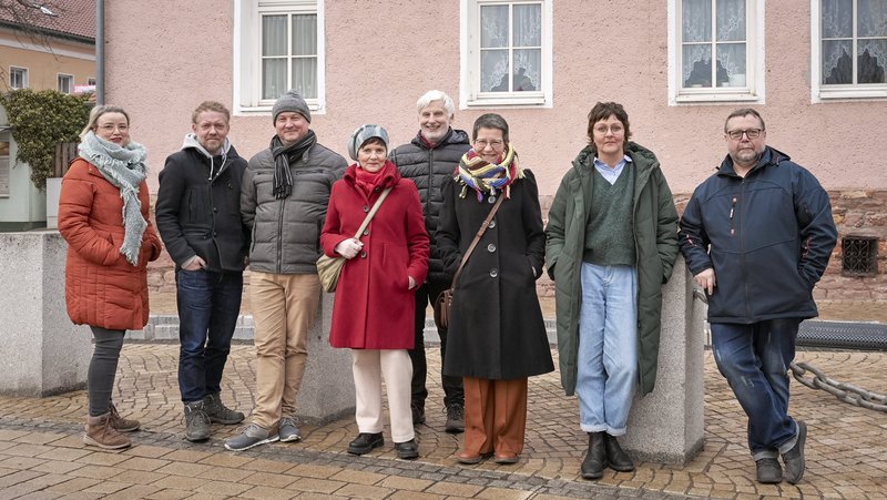 Acht Personen in winterlicher Kleidung stehen locker nebeneinander auf einer Kopfsteinpflasterstraße vor einem rosa Gebäude und lächeln in die Kamera.