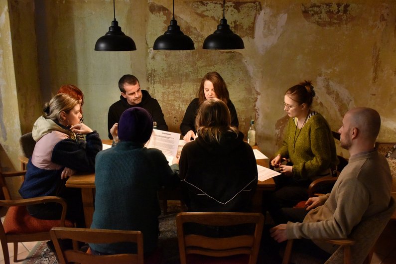 Eine Gruppe sitzt bei Bar-Ambiente um einen Tisch herum, darauf liegen Dokumente