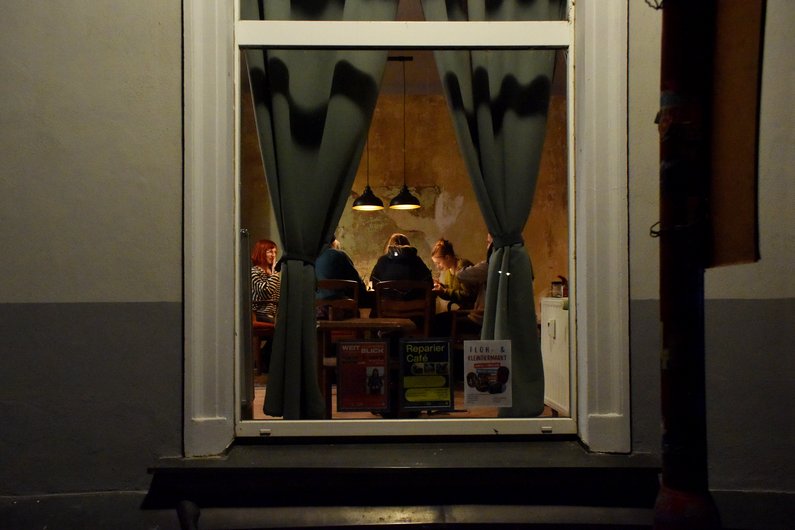 Durch ein Fenster von außen ist eine Gruppe am Tisch sitzend zu sehen, Bar