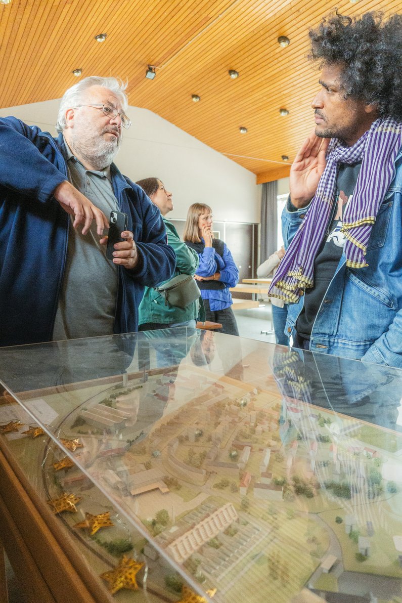 Artist and client stand next to a miniature model, conversing with gestures.