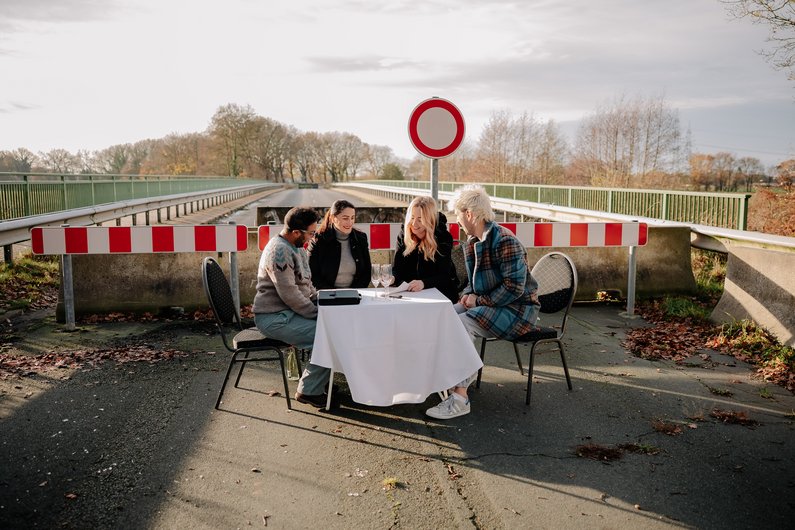 Vier Personen sitzen um einen Tisch im Freien, gedeckt mit Tischdecke und Sekt, vor einer abgesperrten Brücke