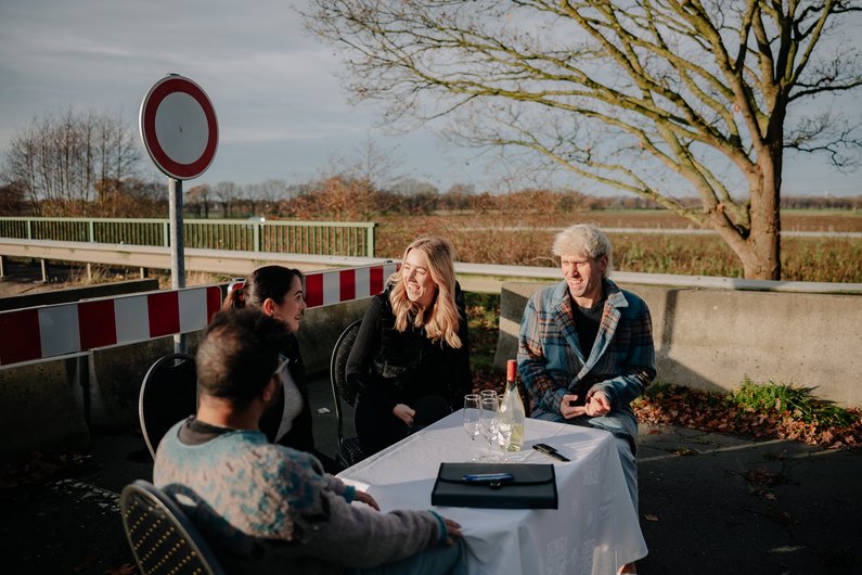 Vier Personen sitzen an einem Tisch im freien, gedeckt mit einer Tischdecke und Sekt, vor einer abgesperrten Brücke