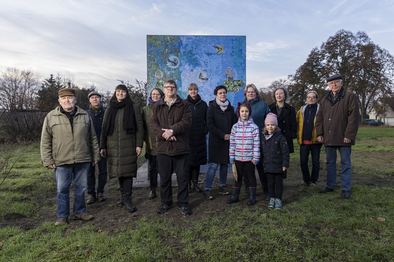 Auftraggebergruppe von Wietstock steht mit Künstlerin Antje Majewski und Mediatorin Susanne Burmester auf einer Wiese vor dem Mosaik