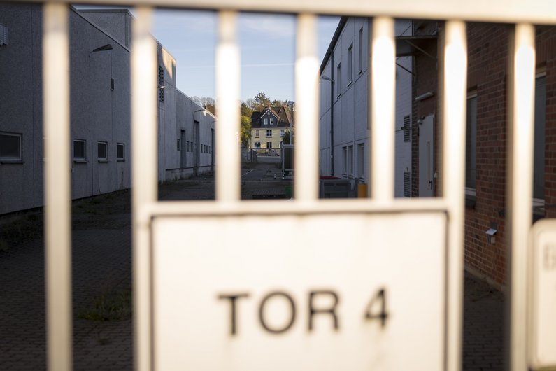 Metallzaun mit einem Schild "Tor 4", dahinter eine Straßenflucht mit niedrigstöckigen  Gebäuden