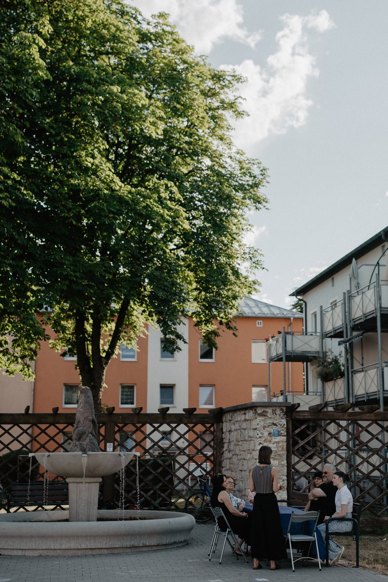 The New Patrons of Amberg – Am Bergsteig View from a distance of a garden table next to a village well, where several people are sitting and talking.
