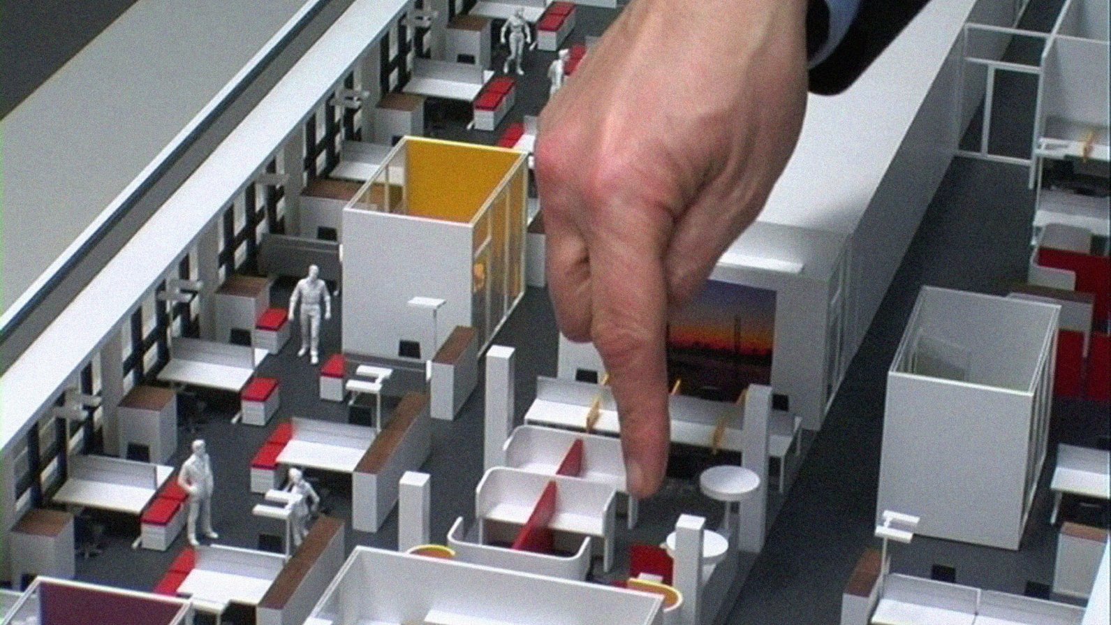 Film still from "A New Product" by Harun Farocki, a hand points to a small element in the interior view of a 3D miniature office model.