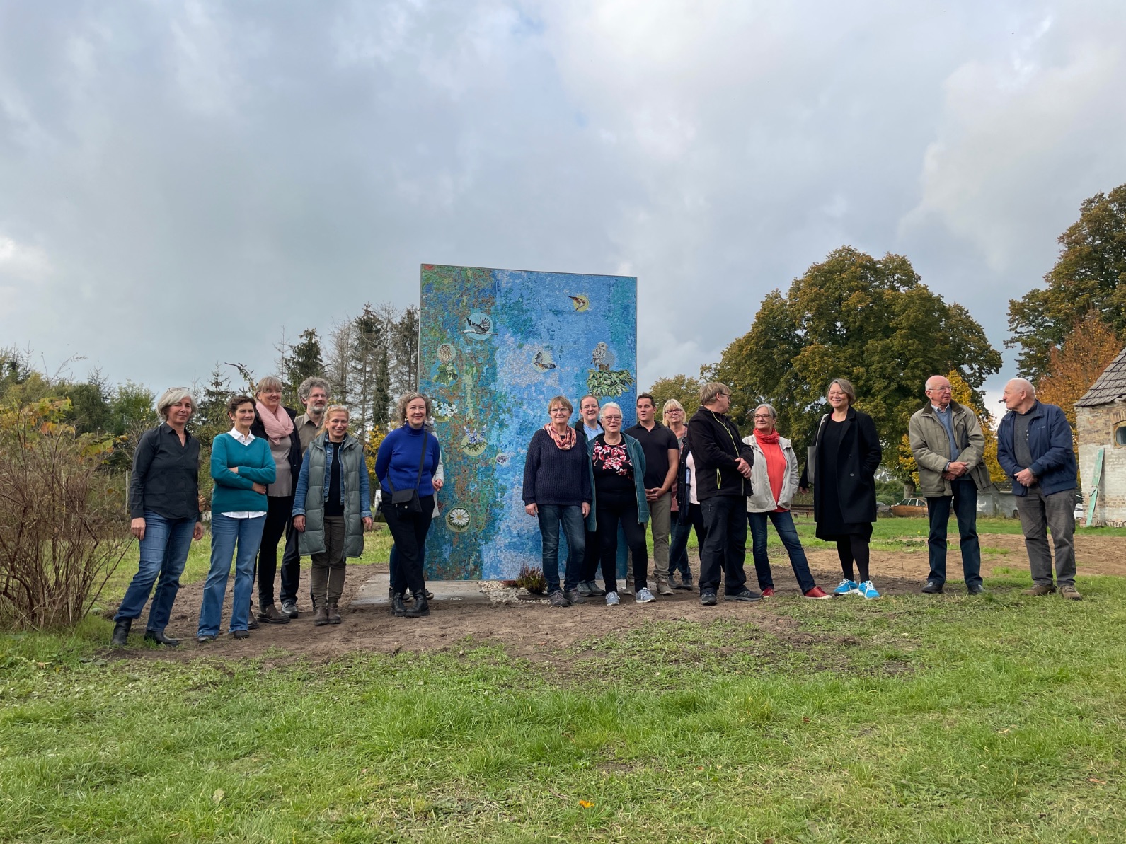 Group photo of The New Patrons' group in front of the mosaic based on the design "Animals and Plants in Wietstock" with artist Antje Majewski and mediator Susanne Burmester 